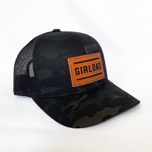 May include: Black and green camouflage trucker hat with a brown leather patch that says "GIRLDAD" in black lettering.