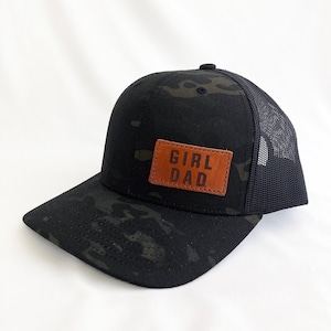 May include: Black and green camouflage trucker hat with a brown leather patch that says "Girl Dad" in black lettering.