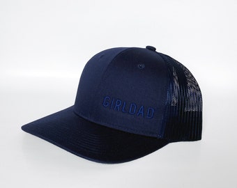 May include: A navy blue trucker hat with a black mesh back. The hat has the word "GIRLDAD" embroidered on the front in blue thread.