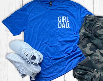 May include: A blue t-shirt with the text "GIRL DAD" in white, paired with white athletic shoes and camouflage print joggers. The items are arranged on a white wooden surface.