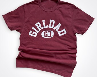 May include: Burgundy t-shirt with white text that reads "Girl Dad" in an arch shape with a white circle containing the letters "GD" in the center.
