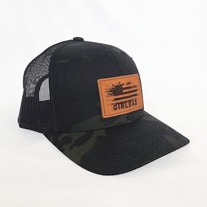 May include: A black and green camouflage trucker hat with a brown leather patch featuring a distressed American flag and the text "Girl Dad".
