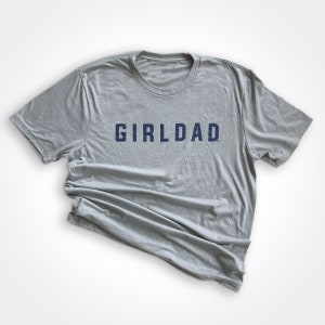 May include: A heather grey t-shirt with the text "GIRLDAD" printed in navy blue.
