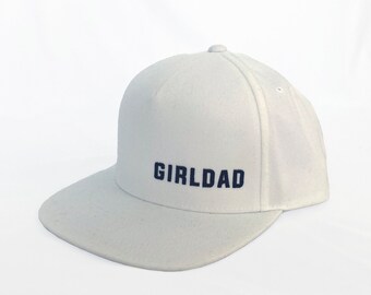 May include: White baseball cap with a navy blue embroidered text that reads "GIRLDAD".