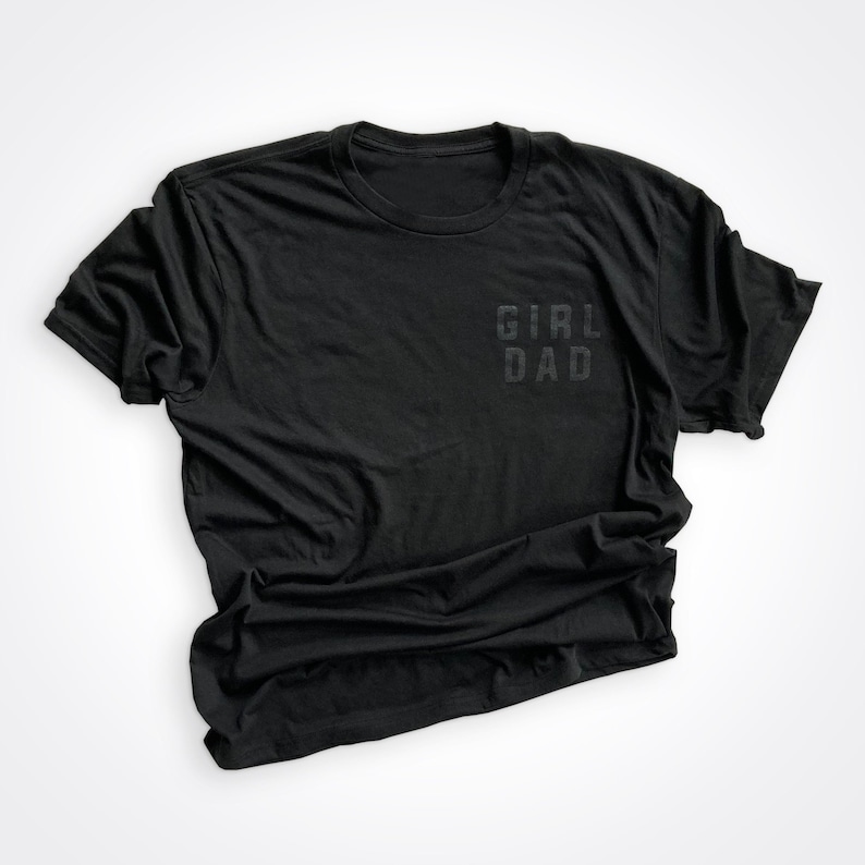 Puede incluir: Camiseta negra con las palabras "Girl Dad" impresas en la parte delantera.