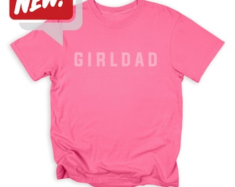 May include: A pink t-shirt with the word "GIRLDAD" printed in light pink across the chest. A red speech bubble with the word "NEW!" in white is in the upper left corner. The shirt is made of a soft material.