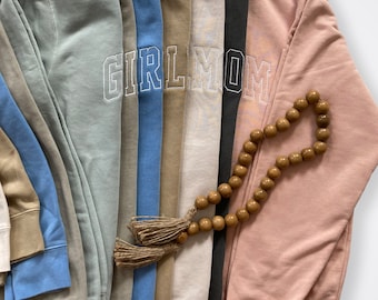 May include: A set of eight sweatshirts in various colors, including light blue, light green, beige, and pink. The sweatshirts feature the text "GIRL MOM" embroidered in white on the front. A wooden beaded necklace is placed on top of the sweatshirts.
