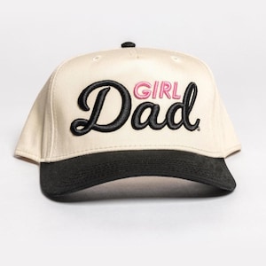 May include: Cream-colored baseball cap with a black brim. The cap features the embroidered text "Girl Dad" in black script with the word "Girl" in pink. The cap has a curved brim and a button on top.