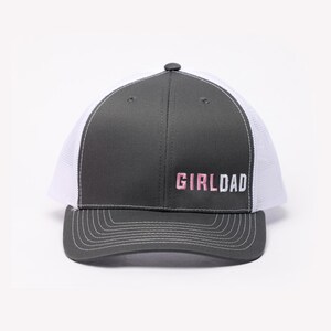 May include: A gray and white trucker hat with the text "GIRLDAD" embroidered in pink and white. The hat features a curved brim, a mesh back, and a snap closure. The hat is a classic style, suitable for casual wear.