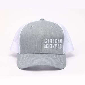 May include: Gray and white trucker hat with the text "GIRLDAD AND BOYDAD" embroidered in white. The front panel is gray, and the mesh back is white. The hat has a curved brim and an adjustable snap closure.