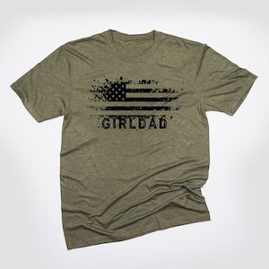 May include: A green t-shirt with a distressed black and white American flag graphic and the text "GIRLDAD" printed on the front.