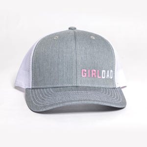 May include: A gray trucker hat with a white mesh back and a curved brim. The hat features the embroidered text "GIRLDAD" in pink and white. The hat is a casual accessory.