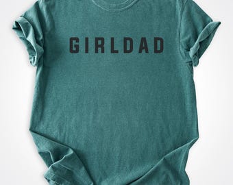 May include: Teal-colored t-shirt with the word "GIRLDAD" printed in black block letters. The shirt has a crew neck and short sleeves. The fabric appears soft and slightly faded.