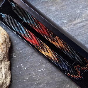 May include: A pair of long, beaded earrings with a geometric pattern in shades of black, blue, red, gold, and yellow.