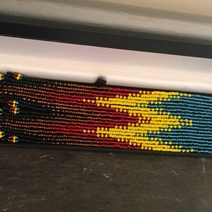 May include: A pair of long beaded earrings in a black box. The earrings are made with black, red, yellow, and blue beads in a geometric pattern.