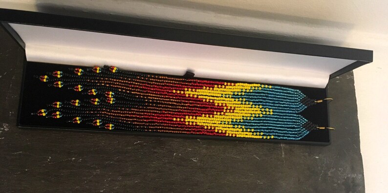 May include: A pair of long beaded earrings in a black box. The earrings are made with black, red, yellow, and blue beads in a geometric pattern.