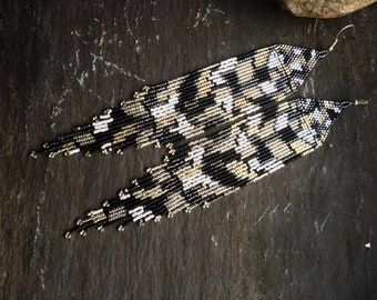 May include: A pair of long, black, white, and gold beaded earrings. The earrings are made with small, round beads that are strung together in a geometric pattern.
