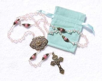 May include: A pink and gold rosary with a silver-toned centerpiece and a cross. The rosary is on a white surface and comes with a light blue velvet pouch.