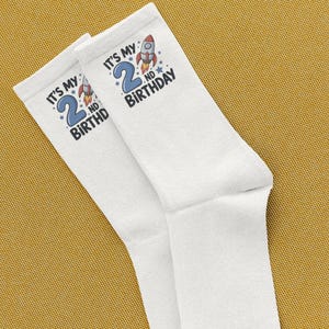 Puede incluir: Un par de calcetines blancos con un diseño de cohete y el texto "IT'S MY 2ND BIRTHDAY" en azul. Los calcetines están hechos de un material suave y están diseñados para el segundo cumpleaños de un niño.