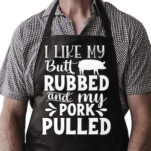 May include: Black apron with white text that reads "I LIKE MY Butt RUBBED and my PORK PULLED" and a pig graphic. The apron has a pocket and is worn over a black and white checkered shirt.