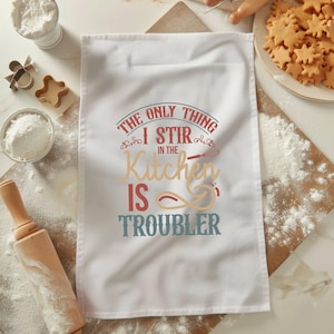 以下が含まれることがあります： 白いキッチンクロスに「The only thing I stir in the kitchen is trouble」という赤い、青い、茶色の文字が書かれています。