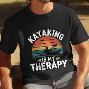 Op de afbeelding: Een zwart T-shirt met een vintage-stijl grafische afbeelding met de tekst "Kayaking is my therapy". De afbeelding toont een zonsondergang met een persoon die kajakt in een kano.