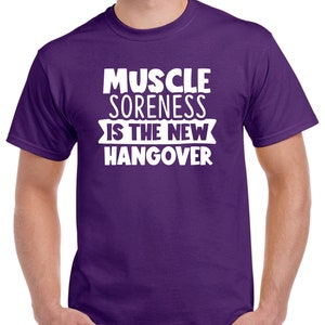 May include: Purple t-shirt with white text that reads "Muscle Soreness Is The New Hangover".