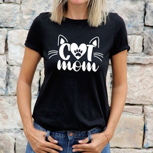 Peut inclure: Un t-shirt noir avec un motif blanc repr&eacute;sentant un visage de chat avec un c&oelig;ur et une empreinte de patte &agrave; l'int&eacute;rieur du c&oelig;ur. Le texte "Cat Mom" est sous le visage du chat.
