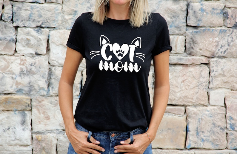 Peut inclure: Un t-shirt noir avec un motif blanc repr&eacute;sentant un visage de chat avec un c&oelig;ur et une empreinte de patte &agrave; l'int&eacute;rieur du c&oelig;ur. Le texte "Cat Mom" est sous le visage du chat.