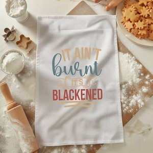 May include: White kitchen towel with the text "It ain't burnt it's blackened" in a vintage style font. The text is in blue, red, and yellow.