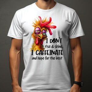 Op de afbeelding: Wit T-shirt met een cartoon-kipillustratie en de tekst "Ik sta niet op en schijn, ik cafeïneer en hoop op het beste."