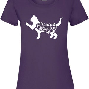 May include: Purple t-shirt with a white silhouette of a cat. The cat's body is formed by the text "All you need is love and a cat".