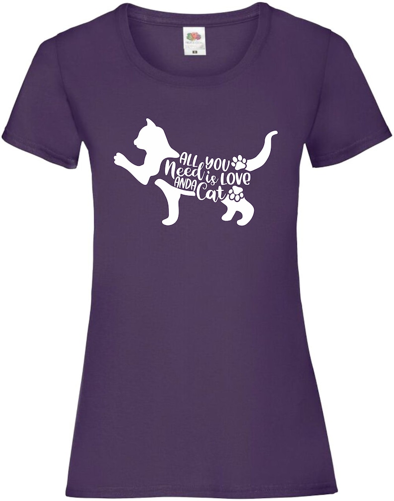 May include: Purple t-shirt with a white silhouette of a cat. The cat's body is formed by the text "All you need is love and a cat".