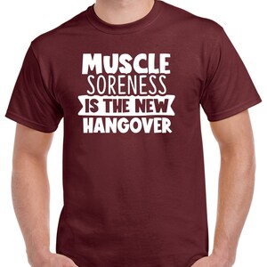 May include: A maroon t-shirt with white text that reads "Muscle Soreness Is The New Hangover".