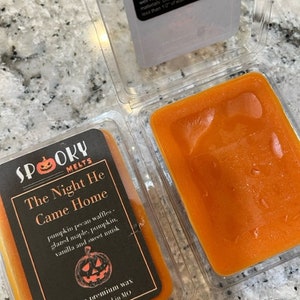 May include: A 2.5 ounce candle in a clear container with an orange wax melt. The label is black with white text and a pumpkin graphic. The text on the label reads "Spooky Melts, The Night He Came Home, pumpkin pecan waffles, glazed maple, pumpkin, vanilla and sweet musk, 2.5oz premium wax, Hand-poured in MO."