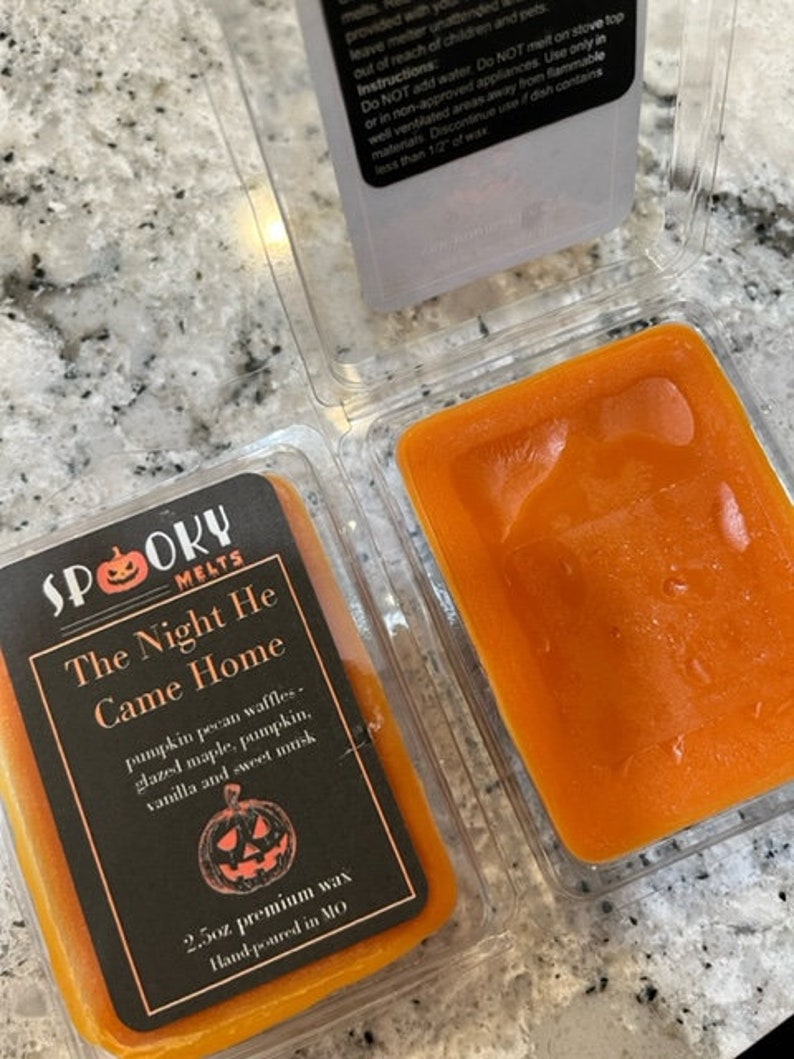 May include: A 2.5 ounce candle in a clear container with an orange wax melt. The label is black with white text and a pumpkin graphic. The text on the label reads "Spooky Melts, The Night He Came Home, pumpkin pecan waffles, glazed maple, pumpkin, vanilla and sweet musk, 2.5oz premium wax, Hand-poured in MO."