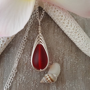 May include: A silver chain necklace with a red glass teardrop pendant wrapped in silver wire. The pendant is on a wooden surface with a seashell.