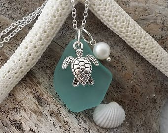 May include: A turquoise sea glass pendant necklace with a silver turtle charm and a white pearl accent. The necklace is displayed on a piece of driftwood with a white seashell.