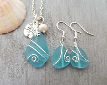 May include: A turquoise sea glass necklace and earrings set with silver wire accents. The necklace features a silver flower charm and a pearl.
