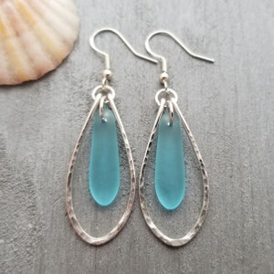 May include: A pair of silver teardrop earrings with a light blue sea glass accent. The earrings have a hammered finish and a simple design.