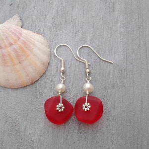May include: A pair of red sea glass earrings with silver accents and white pearl beads. The earrings are on a gray background.