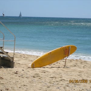 May include: A yellow lifeguard surfboard is lying on the sand on a beach. The surfboard is propped up on a metal stand. The ocean is in the background with two sailboats in the distance. The date and time 04/06/2006 14:33 is visible in the lower right corner of the image.