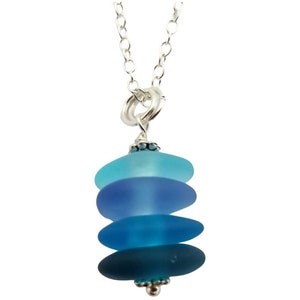 May include: A silver chain necklace with a pendant made of four stacked, graduated, frosted glass ovals in shades of blue. The ovals are stacked from lightest to darkest, with the top oval being a light blue, the second oval being a medium blue, the third oval being a dark blue, and the bottom oval being a teal blue.