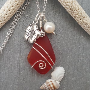 May include: A red sea glass pendant necklace with silver wire wrapping, a silver hibiscus charm, and a white pearl. The necklace is on a silver chain.