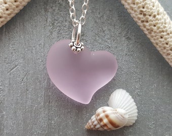 Pode incluir: Um colar com pingente em forma de coração lavanda com corrente de prata. O pingente é feito de vidro fosco e tem uma fiança de prata com um design floral. O colar é exibido com conchas e uma estrela do mar.
