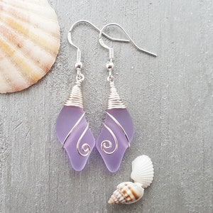 May include: A pair of silver earrings with lavender sea glass wrapped in silver wire. The earrings have a teardrop shape and are dangling.