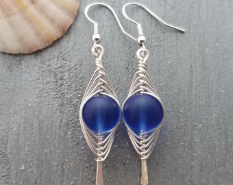 May include: A pair of silver wire wrapped earrings with a dark blue glass bead in each. The earrings are in a diamond shape.
