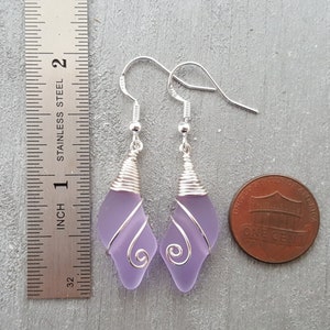 May include: A pair of dangle earrings featuring lavender glass wrapped in silver wire. The earrings are approximately 1 inch long and are shown next to a US penny for size comparison.