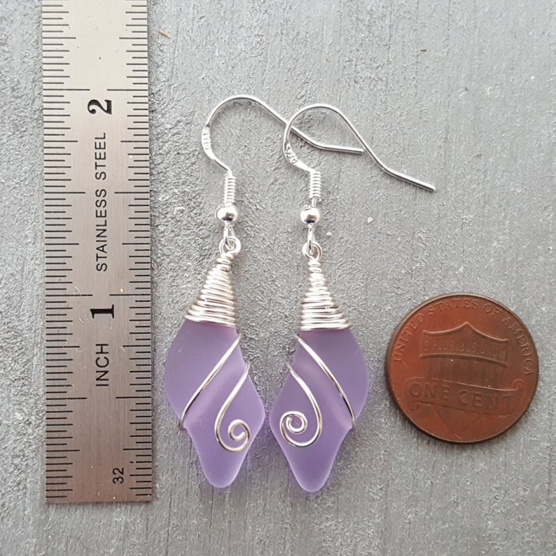 May include: A pair of dangle earrings featuring lavender glass wrapped in silver wire. The earrings are approximately 1 inch long and are shown next to a US penny for size comparison.