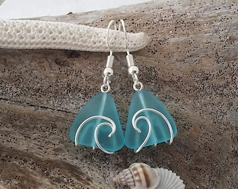 Puede incluir: Pendientes de vidrio de mar azul turquesa con alambre plateado envuelto alrededor del vidrio para crear un diseño de onda. Los pendientes cuelgan de ganchos plateados.